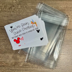 May include: White cards with the text "You've Been Pixie Dusted!" and a Tinkerbell graphic, Mickey Mouse silhouettes, and a red heart. The cards are stacked next to clear plastic bags, all on a wooden surface.