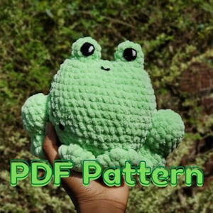 May include: A green crocheted frog plush toy with black eyes. The toy is being held in a hand. The text "PDF Pattern" is visible in the image.