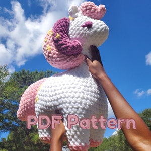 May include: A crocheted unicorn plush toy with a pink and white body, purple and pink mane, and yellow stars. The toy is being held up by a person's hands. The text "PDF Pattern" is visible in the image.