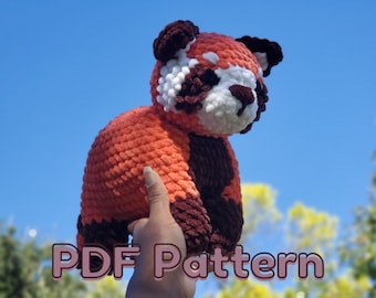 Puede incluir: Un juguete de peluche de panda rojo tejido a crochet con detalles marrones y blancos. El juguete se sostiene en una mano contra un fondo de cielo azul. El texto "PDF Pattern" es visible en la parte inferior de la imagen.