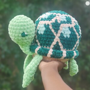 May include: A crocheted green and pink turtle toy with a detailed shell pattern. The turtle is held in a person's hand.