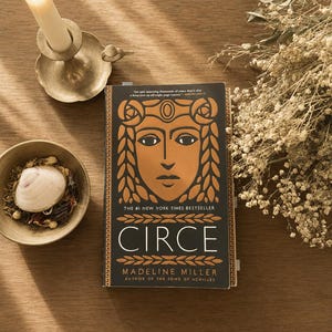 May include: A hardcover book titled Circe by Madeline Miller with a black and gold cover featuring a stylized face illustration. The book is positioned next to a lit candle in a brass holder, a small bowl containing a seashell, and dried flowers.