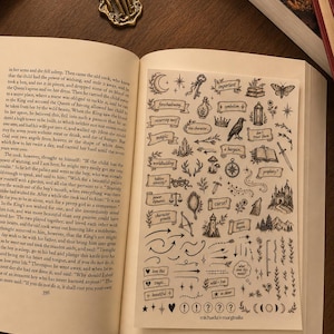 Puede incluir: Una hoja de pegatinas transparente con ilustraciones en tinta negra de varios símbolos, iconos y etiquetas como 'foreshadowing', 'magic' y 'curse', colocada sobre la página de un libro abierto. Incluye dibujos de una luna, llave, corona, cuervo, espada y castillo.
