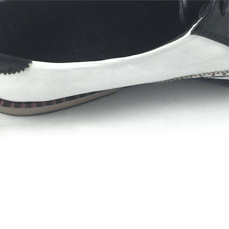 May include: Close-up of the white leather toe of a black and white leather oxford shoe. The shoe has a brown rubber sole.
