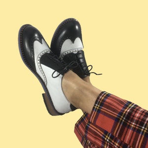 May include: A pair of black and white leather shoes with a wingtip design. The shoes are worn by a person wearing red and black plaid pants.