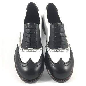 May include: A pair of black and white leather oxford shoes with a wingtip design. The shoes have black laces and a white sole.