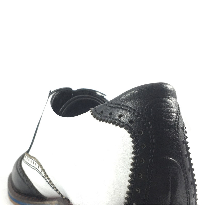 May include: Black and white leather oxford shoes with a wingtip design and a decorative stitch detail. The shoes have a white leather upper and a black leather heel.