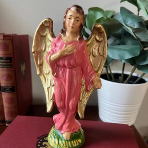 May include: A decorative angel figurine with gold-colored wings and a pink robe. The angel stands on a green and yellow base. The figurine is placed on a red surface, with books and a plant in the background.