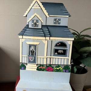 May include: A hand-painted wooden house figurine with a blue and white color scheme. The house has a porch, a window with a welcome sign, and a flower garden. The figurine is on a wooden base.