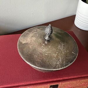May include: A silver bowl with a lid and a pineapple finial. The bowl has a tarnished finish and is sitting on a stack of red books.