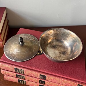 May include: A silver-plated bowl with a lid. The bowl has a slightly hammered finish and the lid has a small knob on top. The bowl and lid are sitting on a stack of red books.