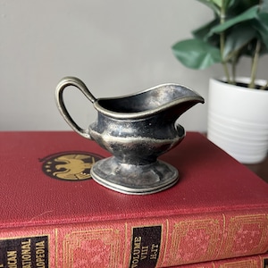 May include: A small, silver-colored creamer or gravy boat with a handle. The boat is sitting on top of a stack of red books with gold lettering.