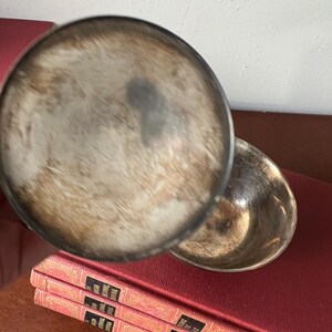 May include: A silver serving dish with a round, shallow bowl and a lid. The lid is slightly ajar, revealing the inside of the bowl. The dish is sitting on top of a stack of red books.