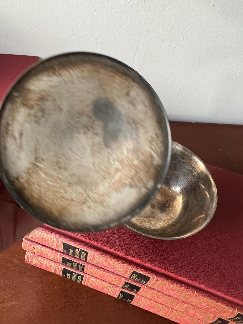 May include: A silver serving dish with a round, shallow bowl and a lid. The lid is slightly ajar, revealing the inside of the bowl. The dish is sitting on top of a stack of red books.