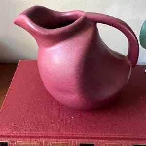 May include: A deep burgundy ceramic pitcher with a unique, asymmetrical shape and a slightly flared lip. The pitcher has a textured surface and a handle that curves up and over the top.