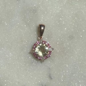 May include: A rose gold pendant featuring a large, round, pale green central gemstone surrounded by a halo of small, round pink stones and four baguette-cut clear stones arranged in a cross pattern, topped with a polished rose gold bail.