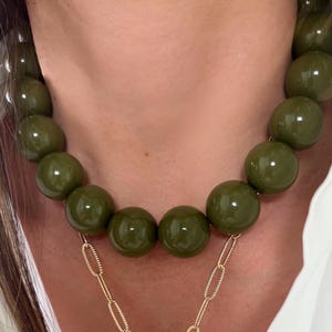 May include: A layered necklace set featuring a short strand of large, glossy olive green spherical beads and a longer gold-tone chain necklace with an elongated oval link design and a central puffed heart-shaped pendant.