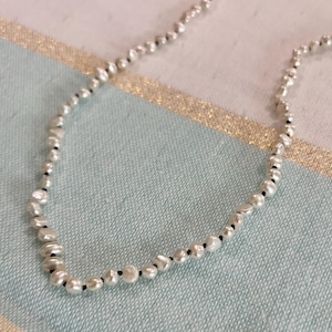 May include: A delicate pearl necklace with irregular, off-white pearls strung on a black cord. The pearls vary in size and shape, creating a unique, organic look. The necklace is displayed on a light blue and gold patterned surface.