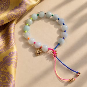 May include: A beaded bracelet featuring smooth, pale green translucent beads strung on a vibrant, multi-colored cord. The bracelet includes a polished gold-tone heart charm and an adjustable sliding knot closure with dangling cord ends finished with small gold-tone beads.