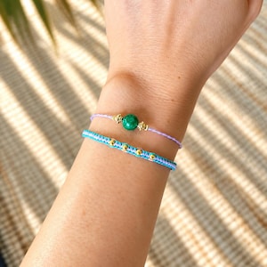May include: Two layered bracelets. The top bracelet is a thin lavender cord featuring a single round green stone bead flanked by two small gold-toned metal accents. The bottom bracelet is a braided turquoise and lavender cord with gold-toned beads.