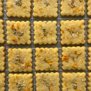 May include: Overhead shot of square-shaped, golden-brown crackers with scalloped edges, arranged on a wire rack. Each cracker is sprinkled with herbs, orange zest, and coarse salt, creating a visually appealing and appetizing display. A close-up view highlights the textures.