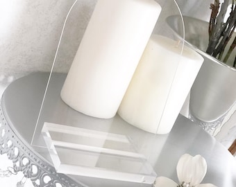 May include: Clear acrylic arch stand with two white pillar candles on a silver cake stand with a white flower.