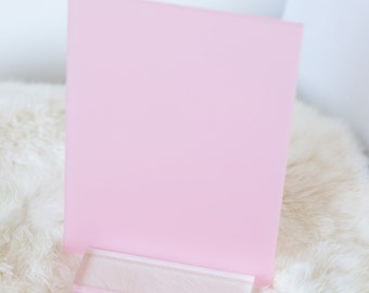 May include: A pink acrylic display stand with a clear acrylic base. The stand is on a white fluffy surface.