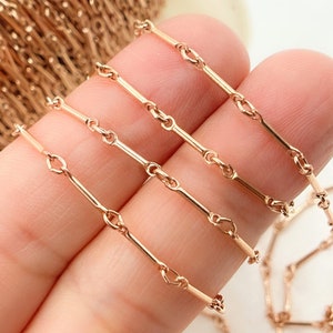 May include: Close-up of a rose gold chain. The chain features a series of elongated, straight links connected by small, heart-shaped links. The chain is held in a person's hand, showcasing its delicate design and warm colour.