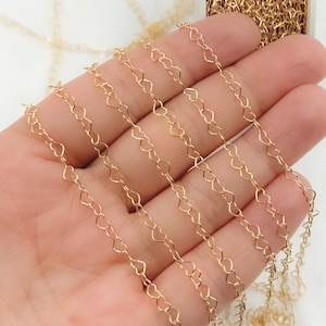 May include: Gold-colored chain featuring a repeating heart design. The delicate chain is displayed against a light background, with a portion of the chain coiled in the upper right corner. This jewelry chain is suitable for crafting necklaces and bracelets.