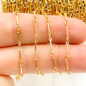 May include: A gold-toned chain featuring a repeating pattern of elongated cylindrical bar links connected by small circular jump rings. The chain is coiled on a spool in the background, with several strands draped across the fingers for display.