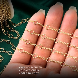 May include: Close-up of a gold-filled clover chain, displayed against a dark green background. The chain features a repeating pattern of diamond-shaped links. Text on the image reads: "14K Gold Filled", "Clover Chain", and "Sold by Foot".