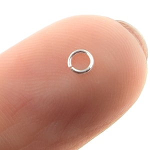 May include: A small, silver jump ring, approximately 5mm in diameter, is shown on a finger.
