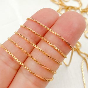 May include: Close-up of several strands of gold-colored box chain necklaces. The chains are displayed against a light background, showcasing their delicate, square-link design. The chains are tightly woven, reflecting light and highlighting their metallic sheen.