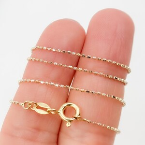 May include: A delicate gold necklace with a beaded chain design. The necklace features a small clasp and is shown against a light background. The chain is made of small, square-shaped beads.