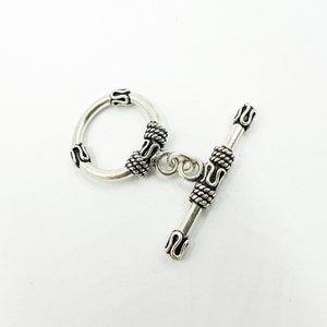 May include: Silver toggle clasp with ornate details. The clasp has a round ring and a bar with a decorative end.