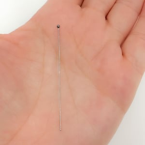 Könnte beinhalten: Eine einzelne silberfarbene Nadel mit einer kleinen, dunkelgrauen Kugel an der Spitze. Die Nadel wird in einer offenen Handfläche vor einem weißen Hintergrund gehalten. Die Nadel ist gerade und dünn.