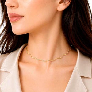 May include: A delicate gold-colored necklace with a wavy design. The necklace is worn around the neck and sits just above the collarbone. The jewelry is a simple, elegant accessory.