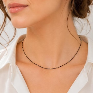 Può includere: Una collana delicata caratterizzata da una sottile catenina nera intervallata da piccole perline color oro distanziate uniformemente. La collana poggia sul collo, mostrando un design minimalista con un aspetto semplice e un profilo sottile e raffinato.