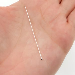 Könnte beinhalten: Nahaufnahme einer silbernen Kopfstiftnadel mit einer kleinen, runden Kugel am Ende. Die Nadel ist dünn und gerade, und die Kugel hat eine glänzende silberne Farbe. Die Nadel wird in einer Hand gehalten.