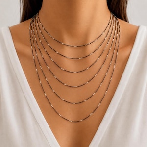 May include: A layered necklace with seven strands. Each strand features a dark chain interspersed with small, silver-colored beads. The necklace is displayed against a white background, creating a contrast that highlights the jewelry.