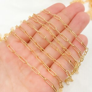 May include: Close-up of a gold-colored chain with an elongated oval link design. The chain is held in a hand, showcasing the intricate details and texture of the links. The chain is likely used for jewelry making.