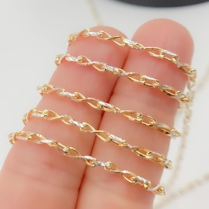 May include: Close-up of a gold and silver chain necklace. The chain has a twisted rope design with alternating gold and silver links. The necklace is held by a person's fingers, showcasing the intricate details and the contrast of the metals.