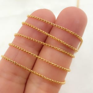 May include: Close-up of a gold chain necklace with a box chain design. The chain is shown in multiple lengths, draped over a finger.