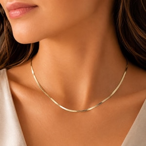 May include: A gold-toned herringbone chain necklace featuring a flat, flexible link design that rests against the collarbone. The necklace has a polished, reflective surface and a continuous, smooth appearance, creating a sleek and uniform look.