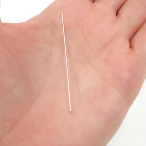 May include: A single, straight, silver-colored head pin. The pin is held in a person's open palm, with the pin's head visible at the bottom. The pin is thin and appears to be made of metal. The background is a light, neutral color.