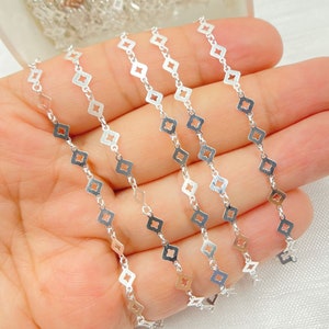 May include: Close-up of several strands of silver-colored chain. Each chain link features a diamond-shaped design with a smaller diamond cut-out in the center. The chains are held in a hand, with a soft, neutral background.