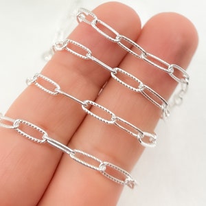May include: Close-up of a silver chain with a textured, oval-shaped link. The chain is shown in a folded position, highlighting the detail of the links.