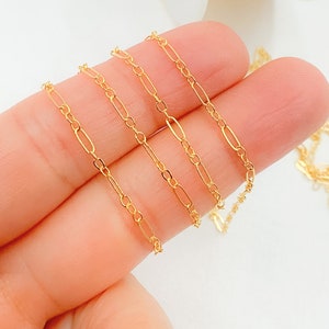 May include: Close-up of a delicate gold-colored chain with an alternating oval and circular link design. The chain is held by a person's fingers, showcasing its fine details and reflective surface. The jewelry is likely for necklaces or bracelets.