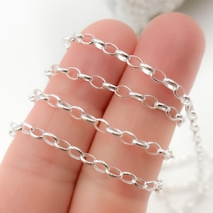 May include: Close-up of a delicate silver chain with oval links. The chain is displayed against a light background, showcasing its reflective, shiny surface. The chain is a jewelry component, suitable for necklaces or bracelets.