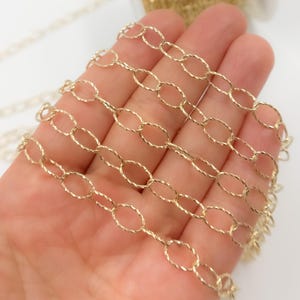 May include: Close-up of a gold-colored chain with oval links. The chain has a textured, twisted design on each link. The chain is held in a hand against a white background. Jewelry making supplies.